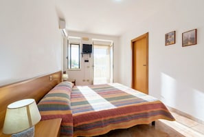 Un dormitorio con cama, lámpara de noche, televisor y ventana.