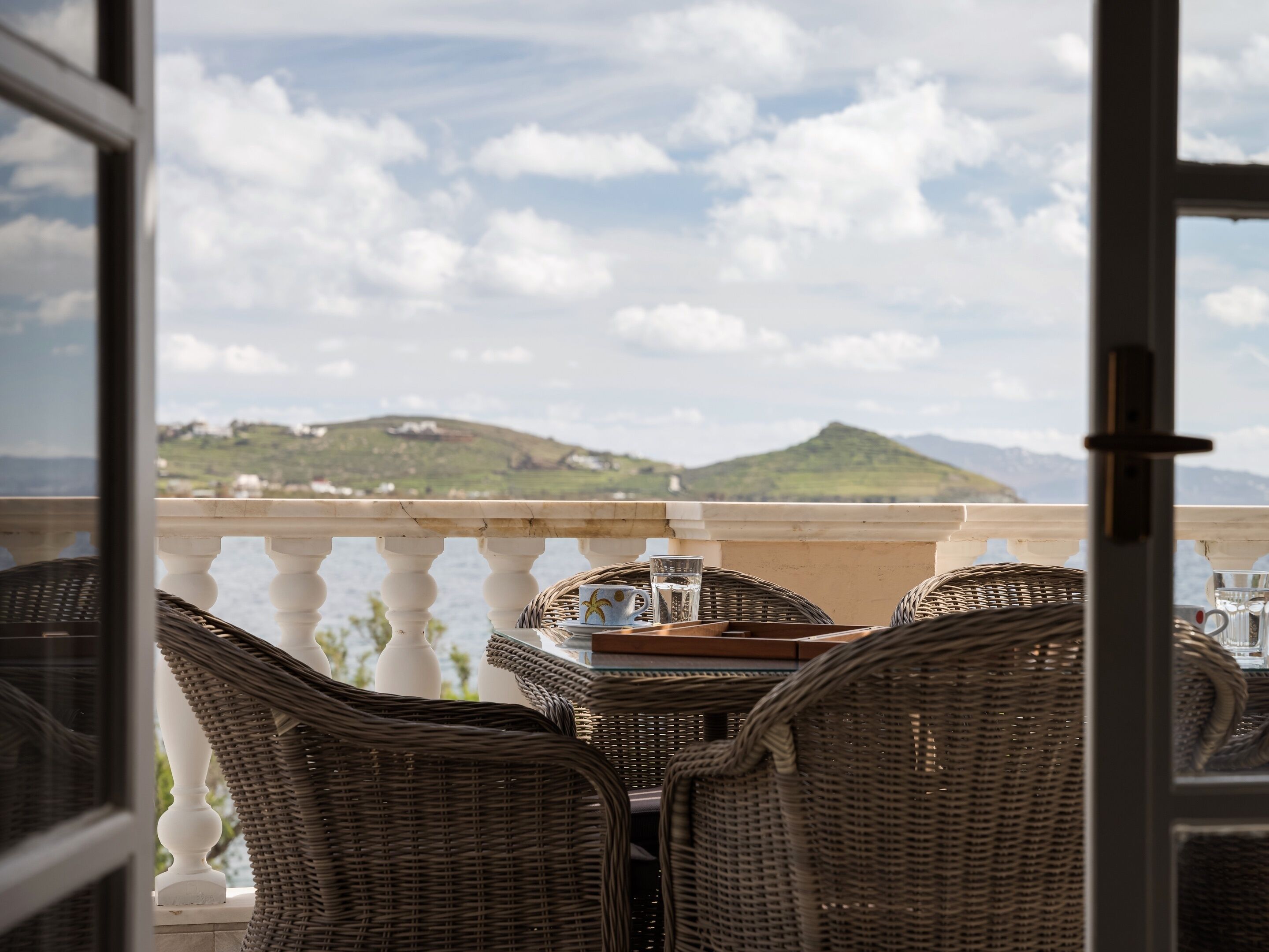 A balcony with wicker chairs and a table, overlooking a scenic view of hills and a body of water.