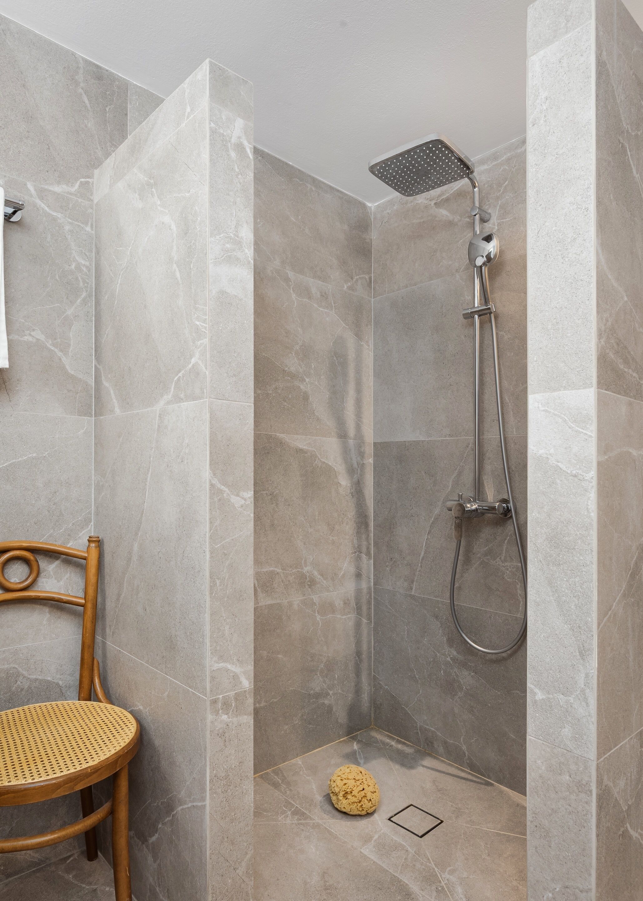 Une douche équipée d’un pommeau de douche à main et d’un pommeau de douche fixé au mur, une chaise en bois et un petit tapis rond sur le sol.