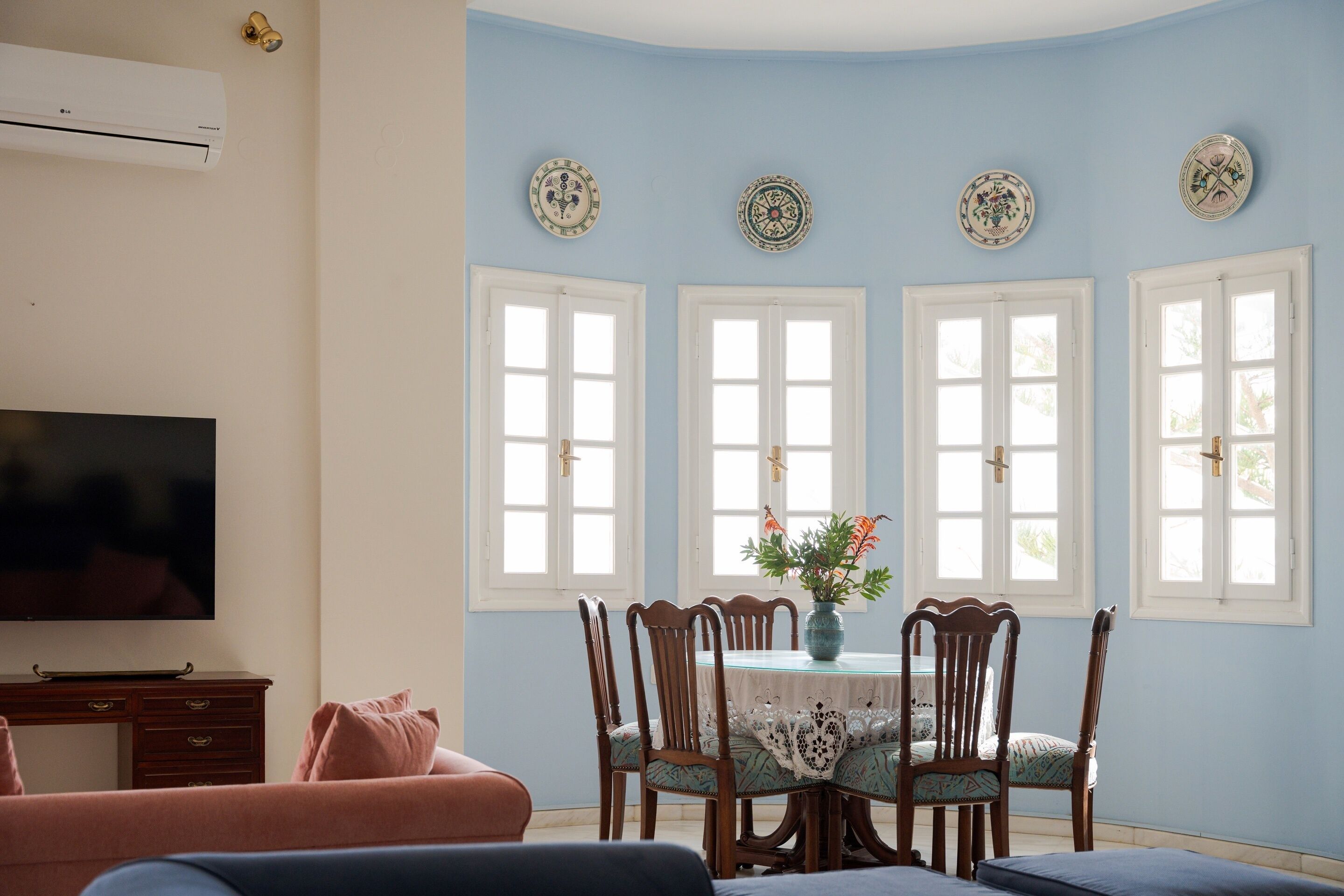 Un coin repas comprenant une table, des chaises et un vase de fleurs. Il y a trois fenêtres à cadres blancs et un téléviseur à écran plat fixé au mur.