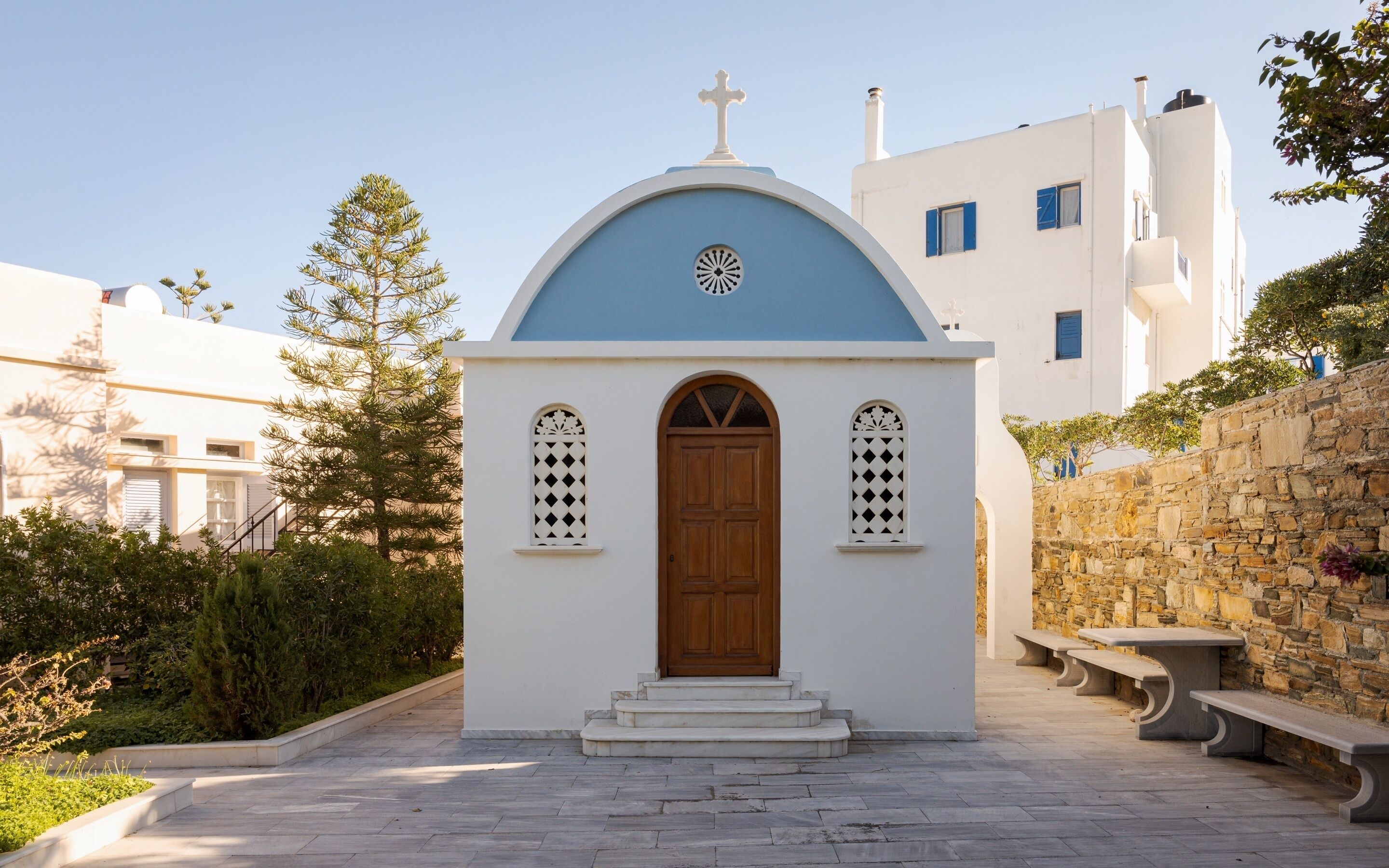 A small white chapel with a blue dome and a wooden door, surrounded by stone walls and greenery.