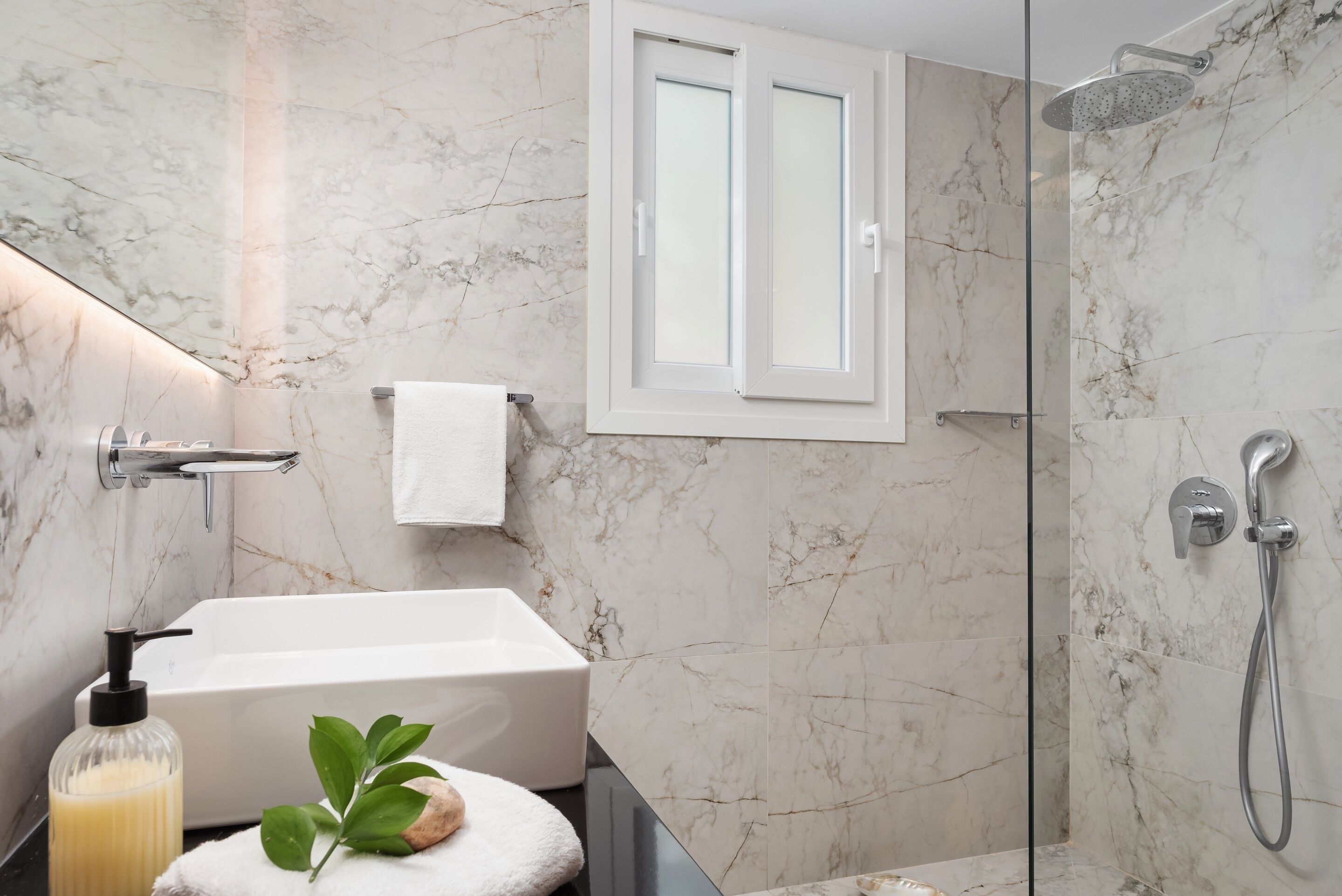 Un baño moderno con pared de mármol, una bañera blanca, cabina de ducha con mampara de cristal y una planta pequeña sobre una toalla.