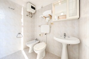Ein modernes Badezimmer mit Toilette, Bidet und Waschbecken, alles in Weiß. Über dem Waschbecken befindet sich ein Spiegel und an der Wand eine Leuchte.