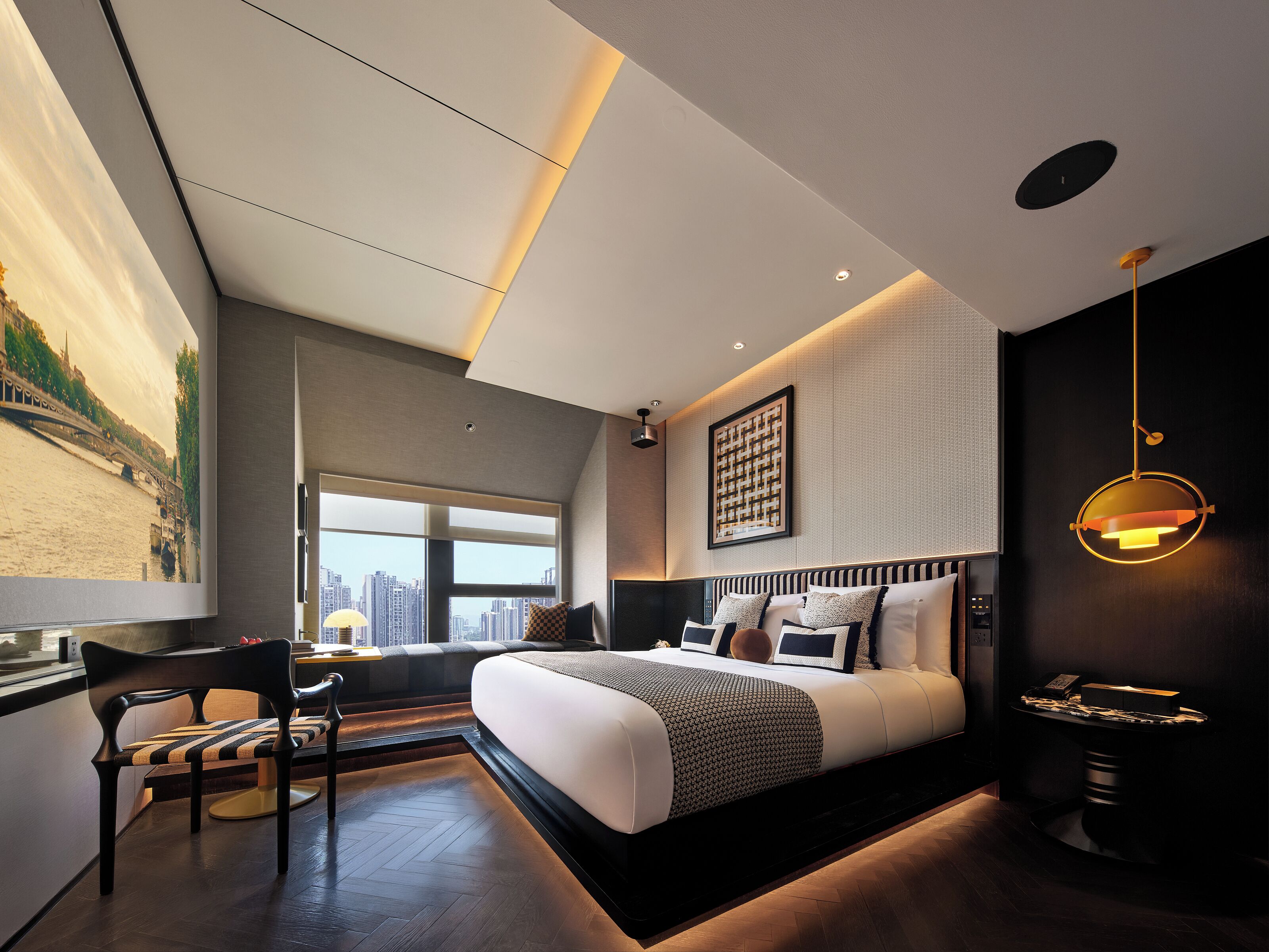 A modern bedroom with a large bed, a desk, and a chair. There is a window with a city view and a painting on the wall.
