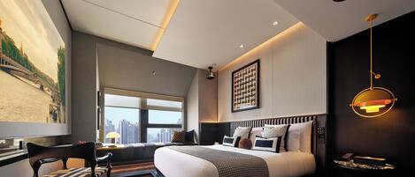 A modern bedroom with a large bed, a desk, and a chair. There is a window with a city view and a painting on the wall.