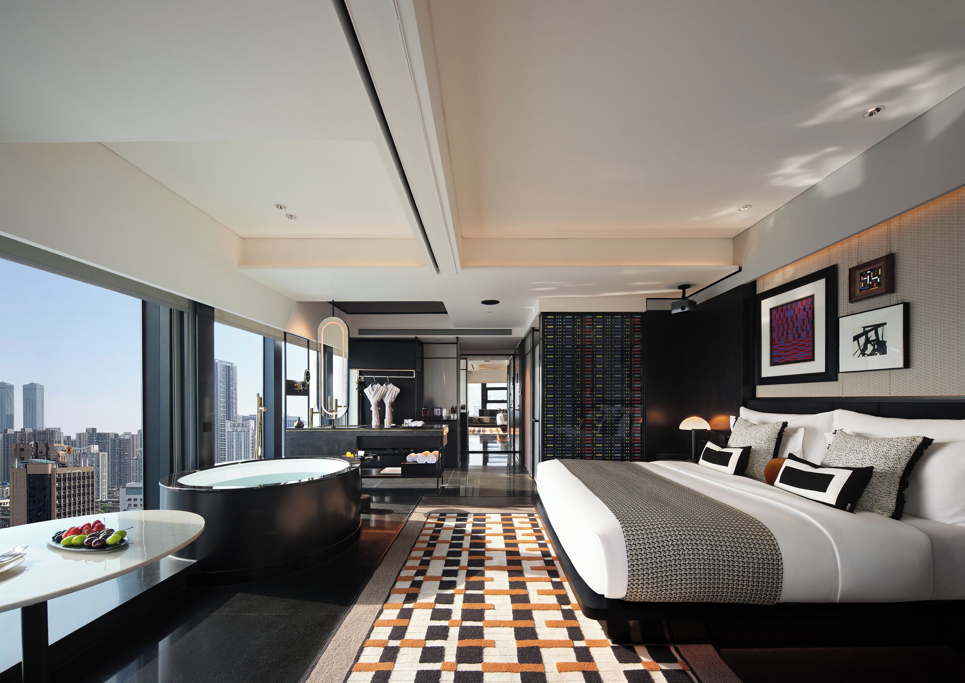 A modern hotel room with a large bed, a sitting area with a coffee table, a bathtub, and a city view.