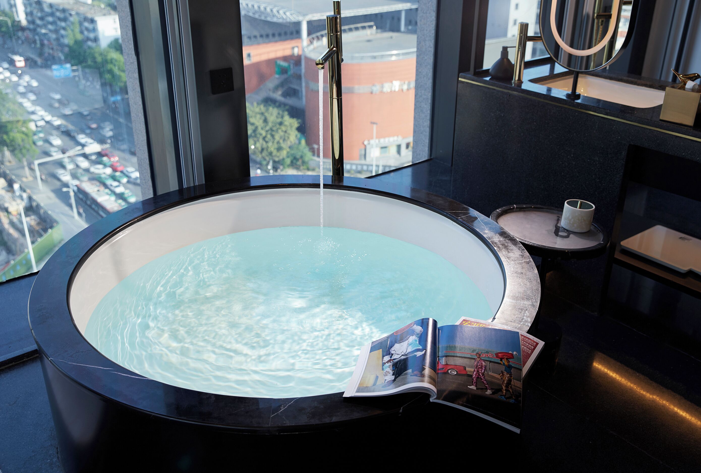Un baño moderno con una bañera grande, una ventana con vista a la ciudad y una revista al borde de la bañera.