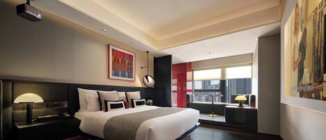 A modern hotel room with a large bed, a ceiling projector, a city view, and a framed artwork on the wall.