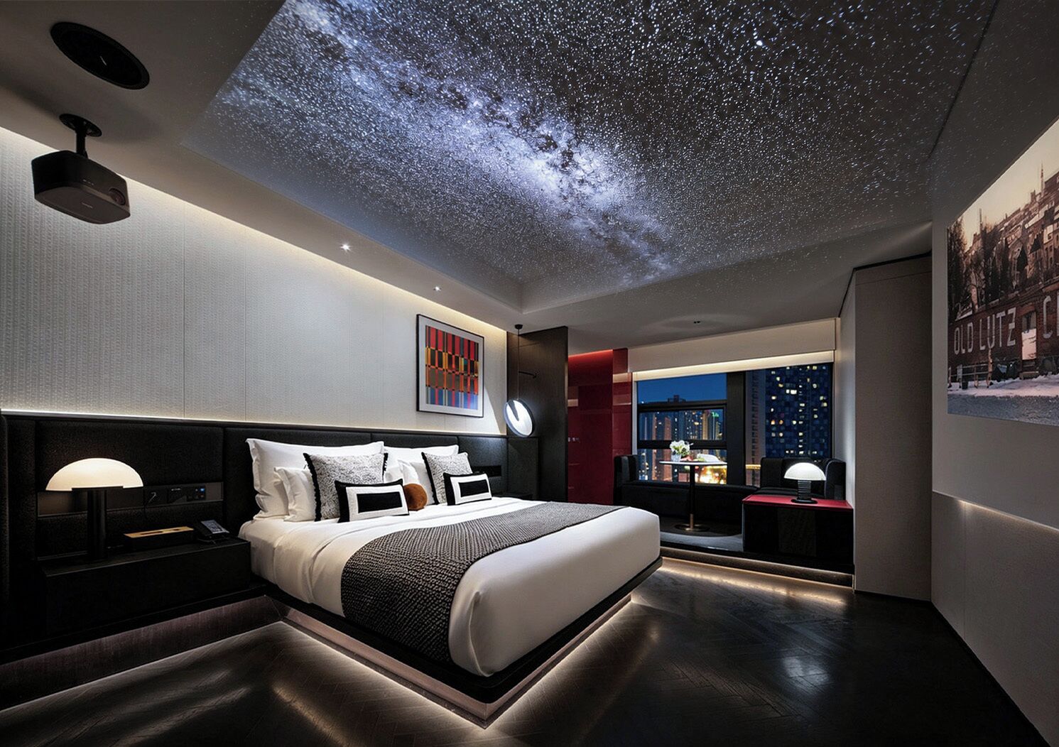 Une chambre moderne avec un lit, des tables de chevet, une petite table, un projecteur au plafond qui diffuse une image de ciel étoilé, une vue sur la ville à travers une grande fenêtre et un tableau encadré au mur.