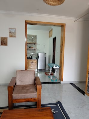 Un salon avec un fauteuil en bois et une table basse, un coin cuisine équipé d’un réfrigérateur et d’une bouilloire, et une porte donnant sur une autre pièce.