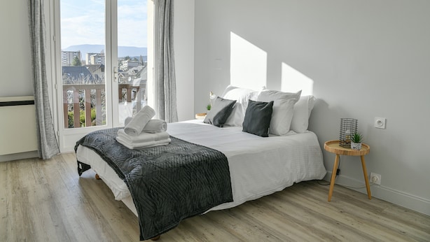 Un dormitorio con cama, una mesita de noche, una ventana con cortinas y vistas a edificios.