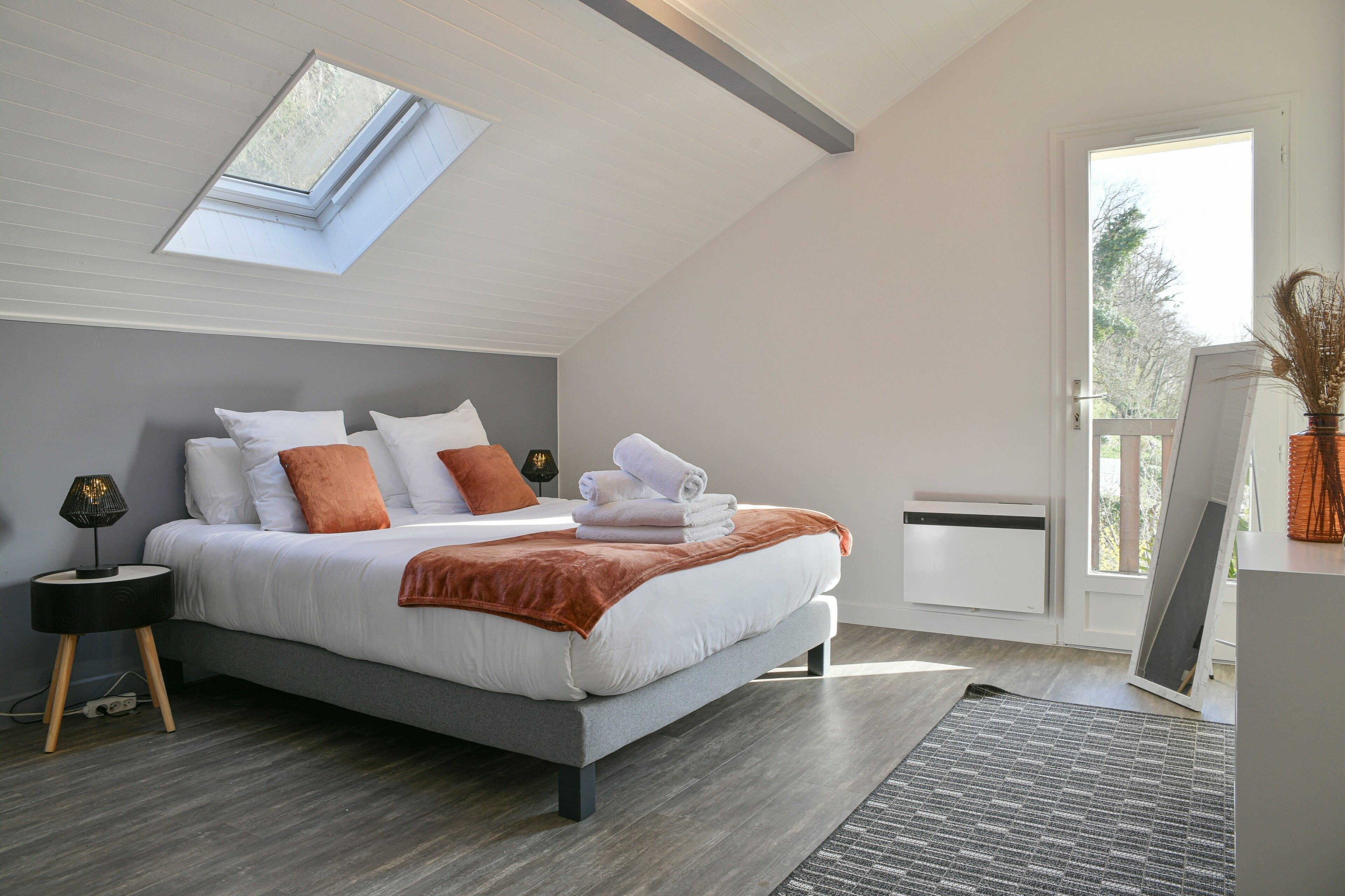 A bedroom with a skylight, a bed with pillows, a bedside table with a lamp, and a view through a door.