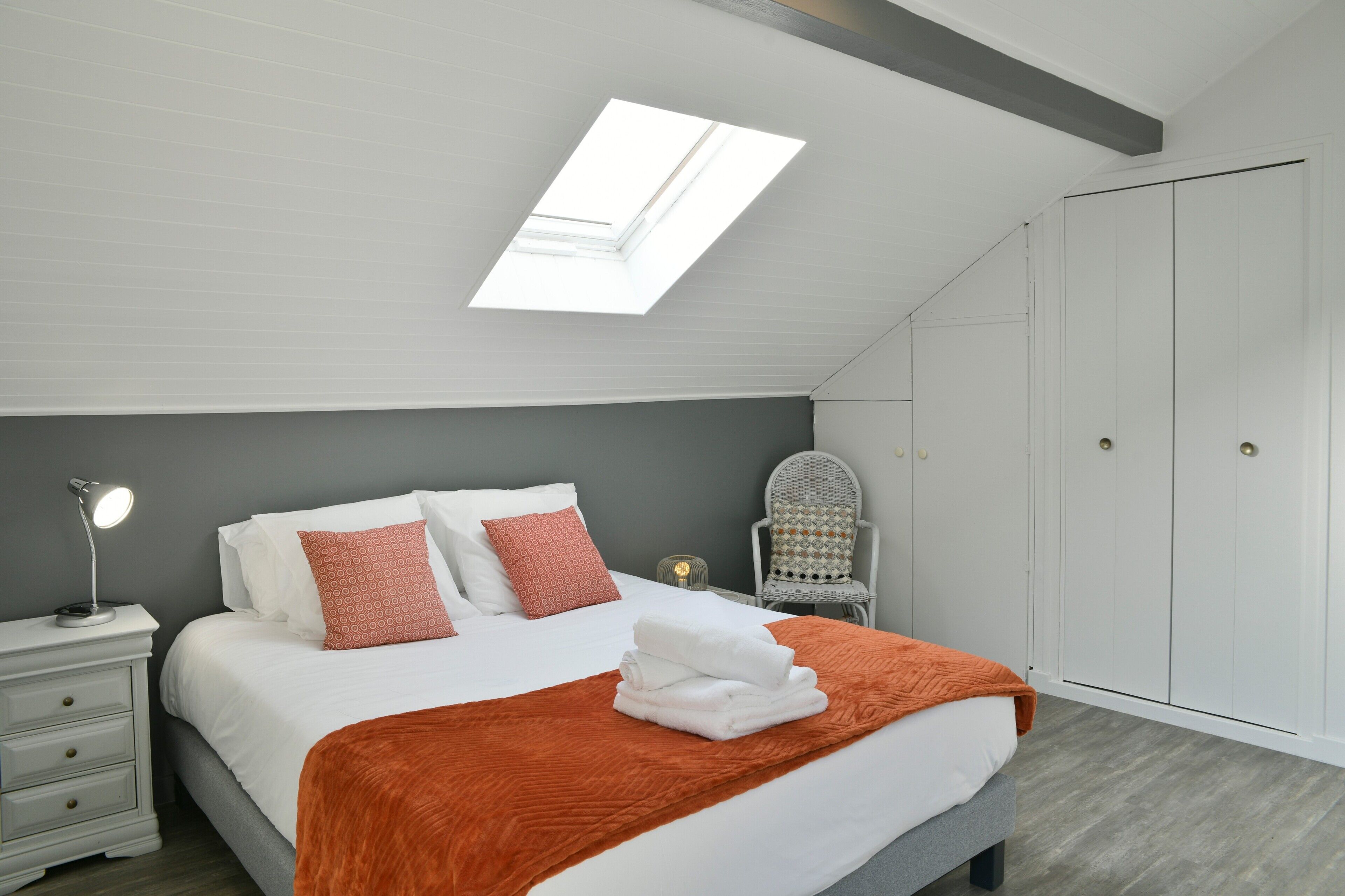 Ein Schlafzimmer mit Dachfenster, einem Bett mit weißer Bettwäsche, einer orangefarbenen Tagesdecke, einem Nachttisch mit Lampe und einem Kleiderschrank.