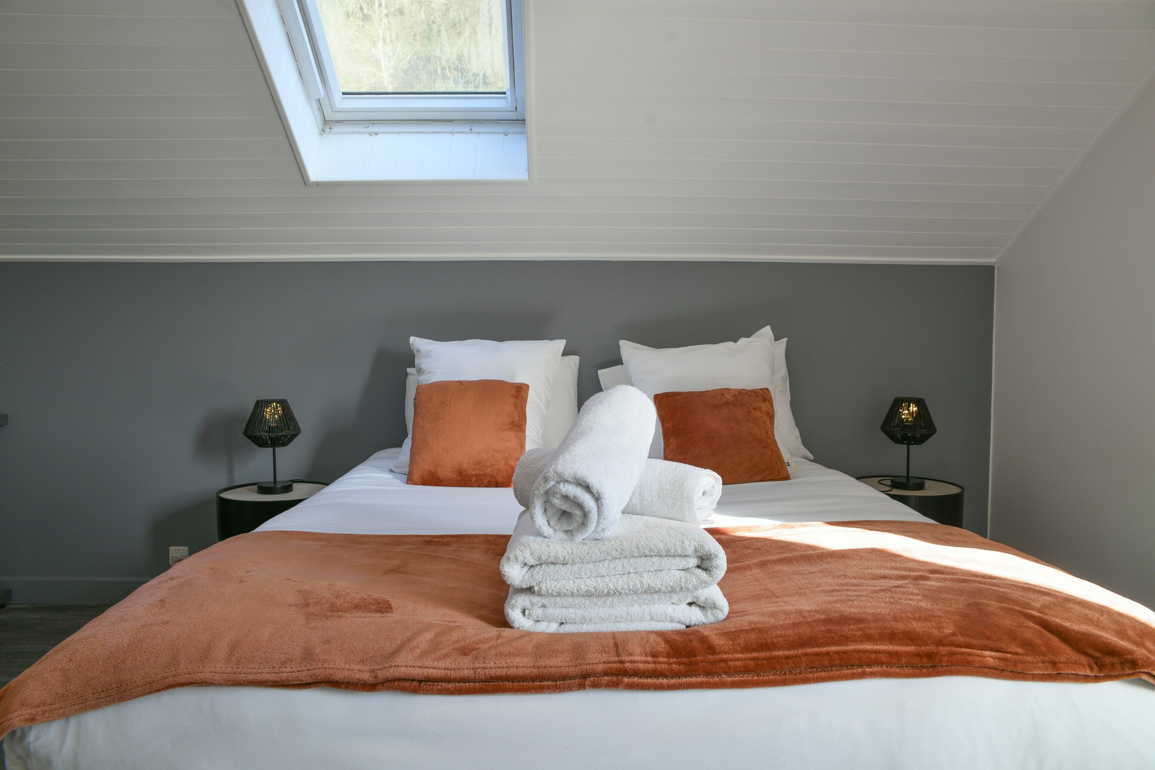 A neatly made bed with a brown blanket, white pillows, and a rolled towel on top. There are two bedside lamps on either side of the bed.
