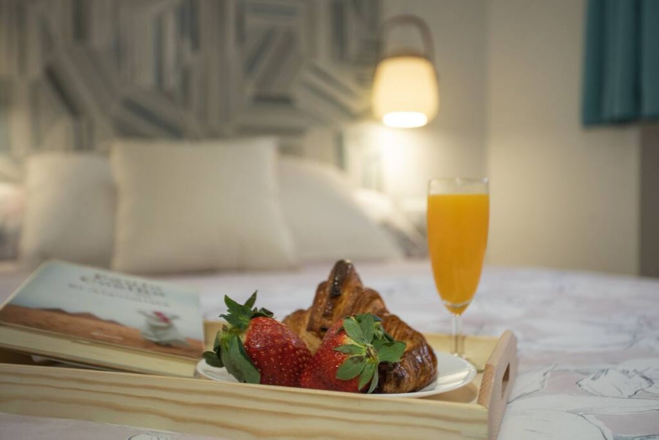 Un plateau de petit-déjeuner avec des croissants, des fraises et un verre de jus d’orange, posé sur un lit avec un livre et des oreillers en arrière-plan.