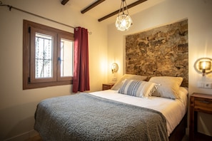 A bedroom with a stone wall, a bed with a grey blanket, a wooden headboard, a hanging light fixture, and a window with curtains.