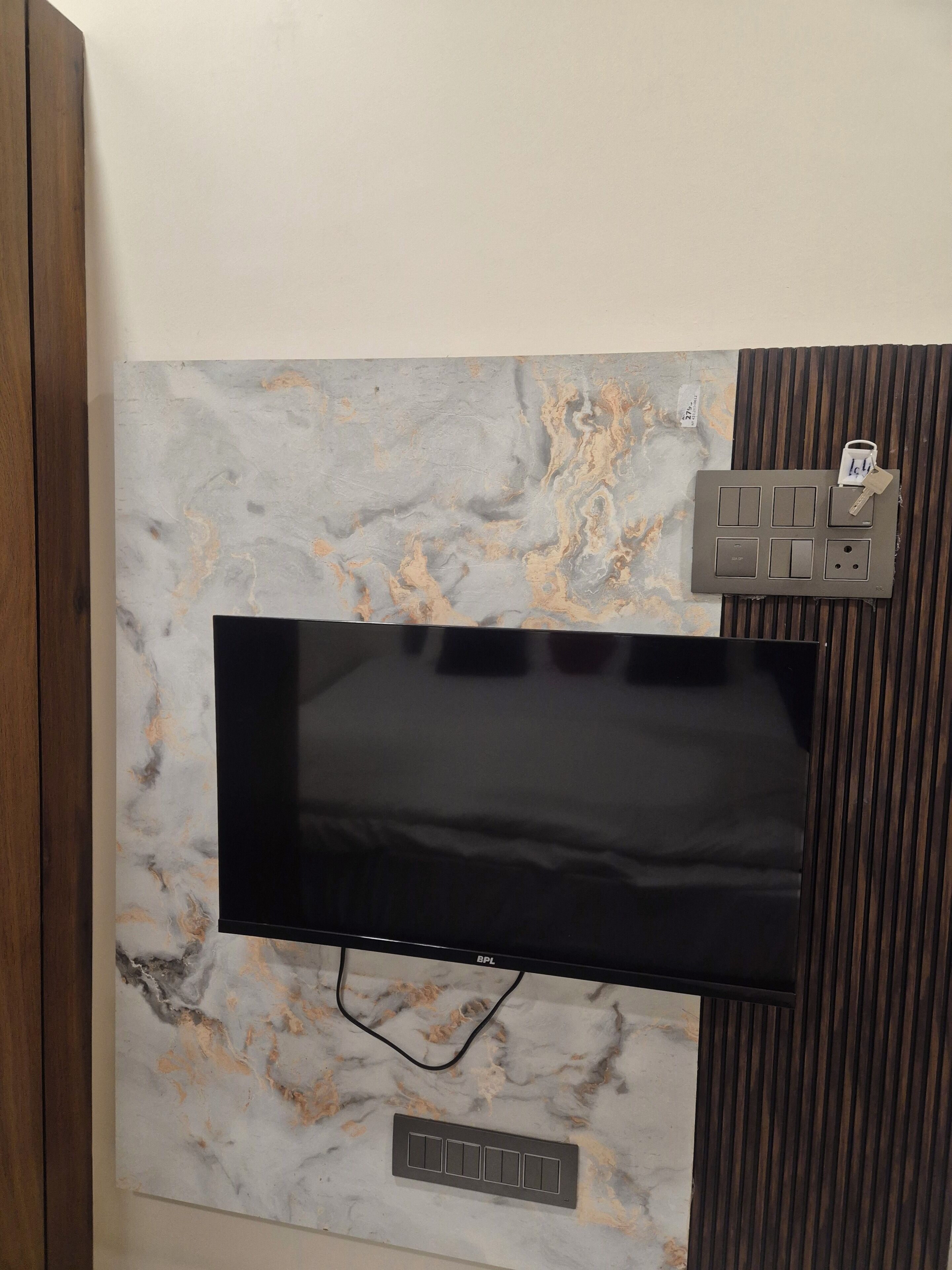 A flat-screen TV mounted on a wall with a marble-like pattern, a power outlet, and a switchboard.