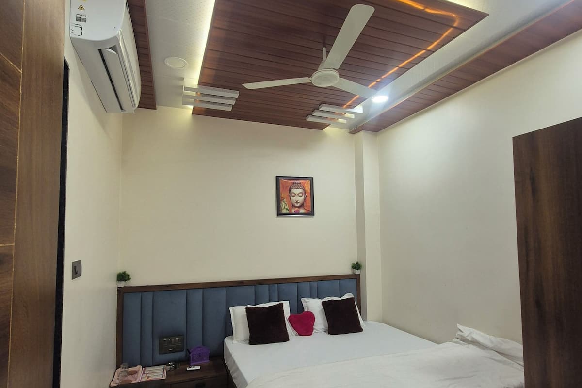 A bedroom with a bed, a ceiling fan, an air conditioning unit, a nightstand, and a small plant.
