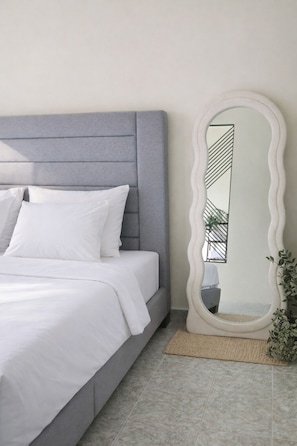 A bedroom with a grey upholstered headboard, white bedding, and a large, white, wavy-framed mirror on the wall.