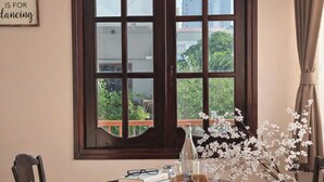 A dining area with a round table, two chairs, a decorative hanging light, and a large window with a view of buildings and greenery.