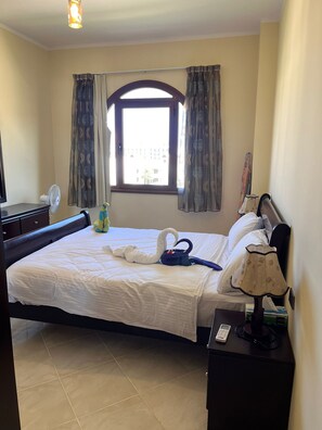 Una habitación de hotel con una cama, una mesita de noche con lámpara, un ventilador y una ventana con cortinas.