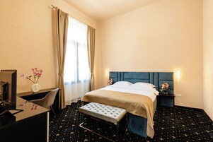 Habitación de hotel con cama, escritorio, silla, una pequeña mesa con un florero y mesita de noche con lámpara.