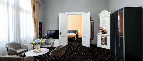 Quarto de hotel com cama, televisão, mesa de jantar com cadeiras, pequena cozinha e uma grande janela com cortinas.