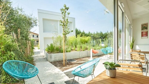 Un espace patio avec une table et des chaises en bois, entouré de verdure et avec un bâtiment blanc en arrière-plan.