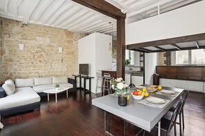 A modern living room with a stone wall, a sofa, a dining table set with plates and fruit, and a kitchen in the background. - Spacious 2BR in The Heart of Saint Germain (Paris)