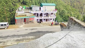 A two-story building with a green roof and multiple balconies, located near a road with a guardrail and a rocky hillside in the background. - Hotel Shri Ganesh Hari Ganesh (Ukhimath)