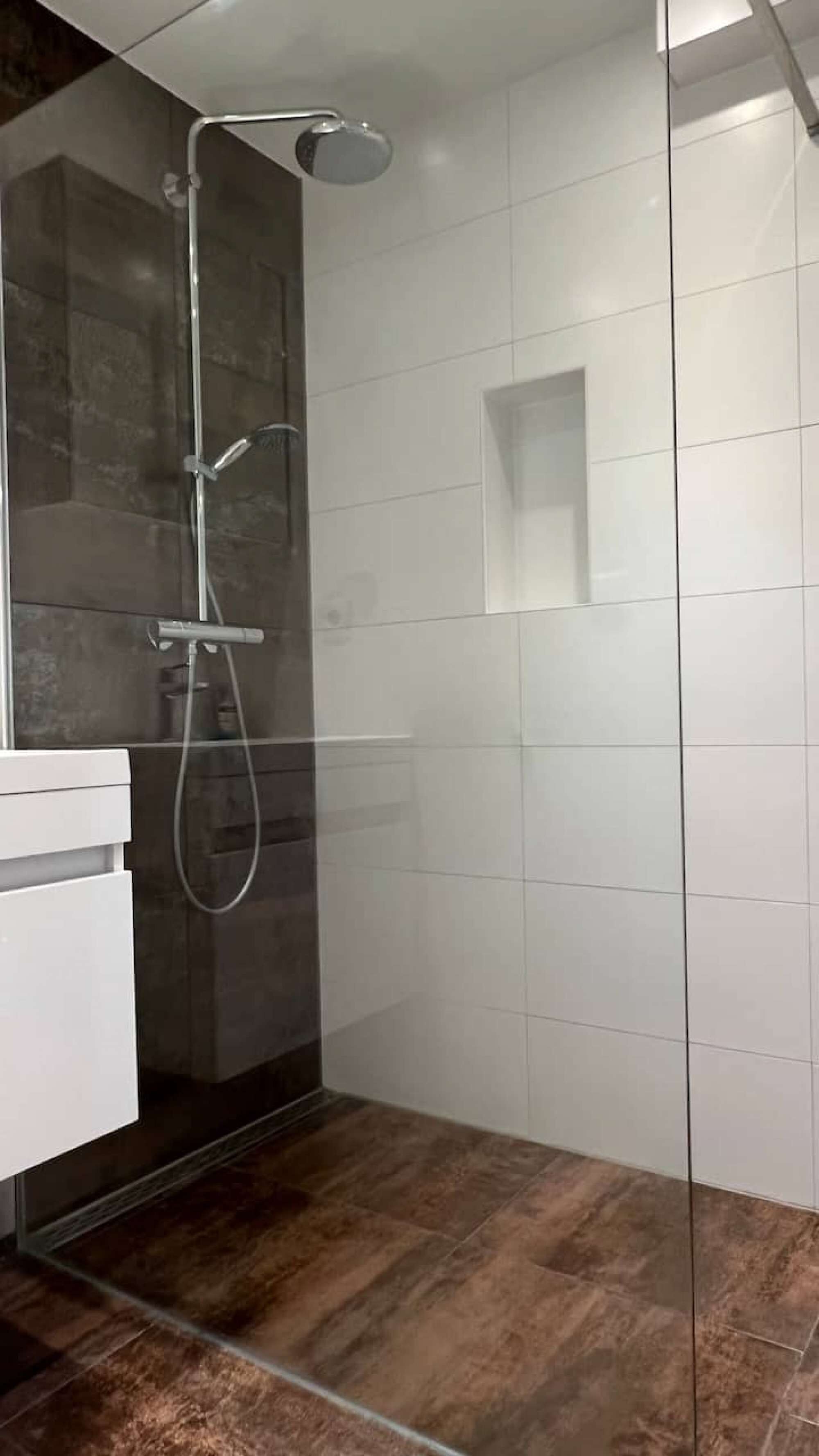 Un baño moderno con ducha de vidrio, lavamanos blanco y suelo de baldosas oscuras.