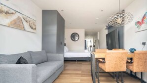 Ein modernes Hotelzimmer mit grauem Sofa, einem Holzespeisetisch mit Stühlen, einem Bett und einem großen Fenster.
