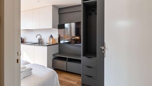 Et kompakt, moderne værelse med tekøkken, en seng med pænt foldede hvide håndklæder og en grå garderobe med flere skuffer.