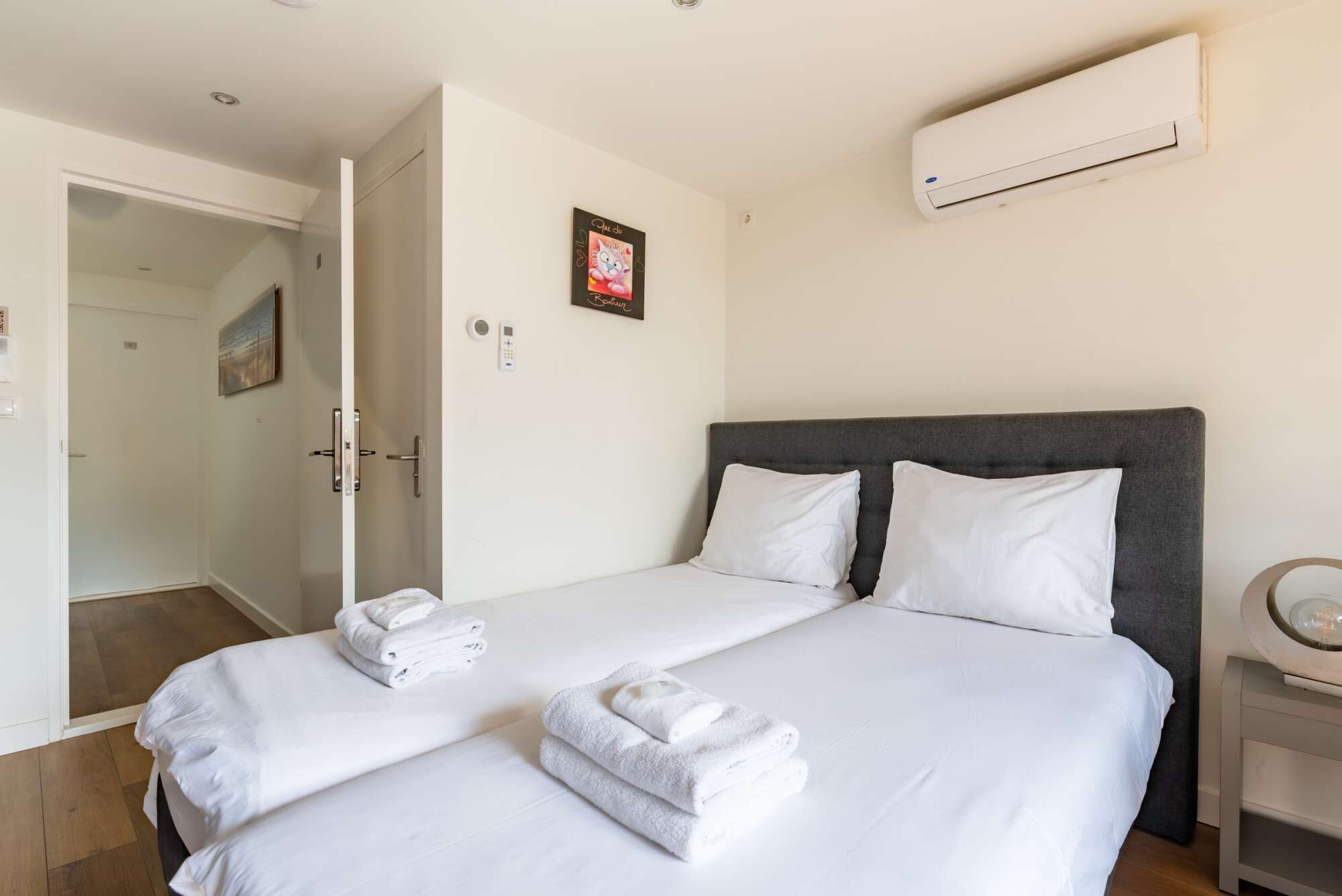 A hotel room with two beds, white linens, a gray headboard, an air conditioning unit, a door with a glass panel, and a small table with a lamp.