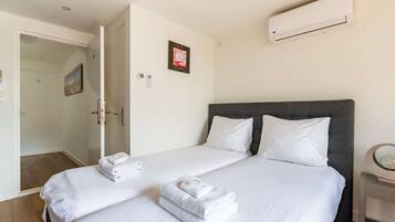 Habitació d'hotel amb dos llits, roba blanca, un capçal de llit gris, un aparell d'aire condicionat, una porta amb un panell de vidre i una taula petita amb una làmpada.