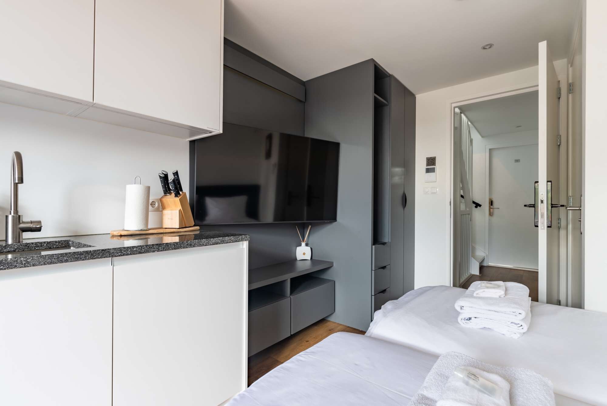 Ett modernt hotellrum med ett litet kök, en platt-TV, en säng med prydligt vikta handdukar och en rymlig klädkammare.