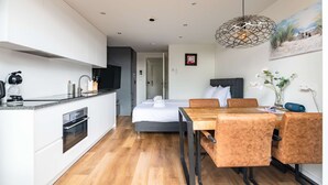 Et moderne hotelværelse med et tekøkken, et spiseområde med et træbord og stole, en seng med hvide sengetøj og en unik geometrisk pendel.