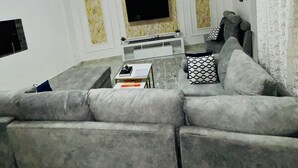A modern living room with a grey sectional sofa, a flat-screen TV mounted on a marble wall, a small white coffee table, and a ceiling light fixture.