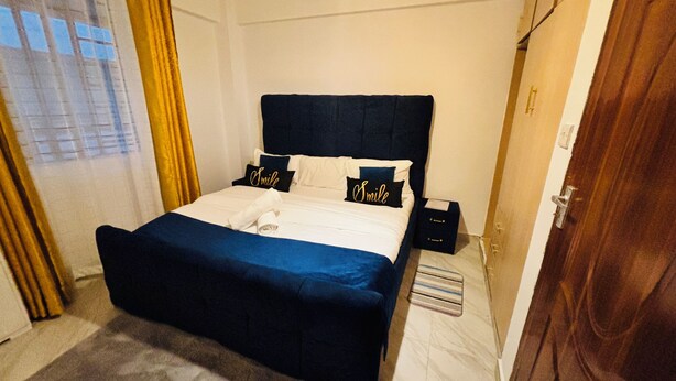 A neatly made bed with a dark headboard, two pillows with the word "Smile," a small bedside table, and a window with curtains.