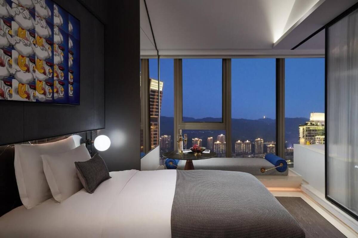 Ein modernes Hotelzimmer mit einem großen Bett, einem runden Tisch und Blick auf die Stadt durch große Fenster.