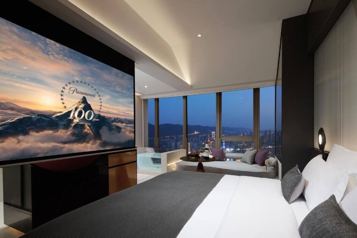 Ein modernes Hotelzimmer mit großem Fernseher, der ein Paramount 100 Logo zeigt, einem großen Bett, einer Sitzecke mit Blick auf die Stadt und einem großen Fenster.