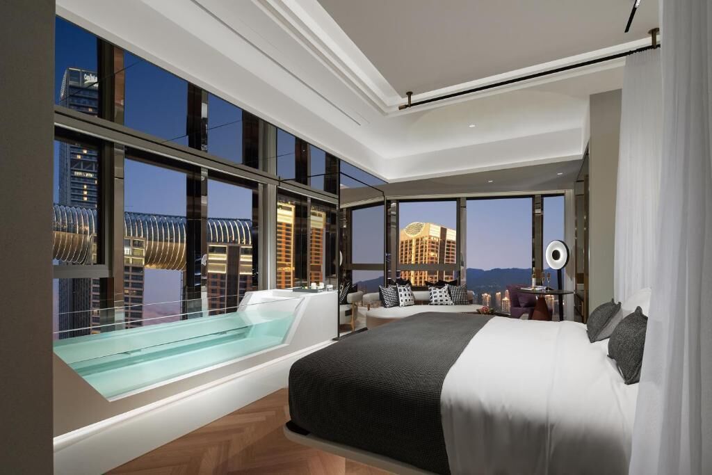 A modern hotel room with a large bed, a glass-enclosed jacuzzi, and a city view.