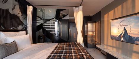 A modern hotel room with a large bed, a view of a staircase, and a Paramount movie poster on the wall.