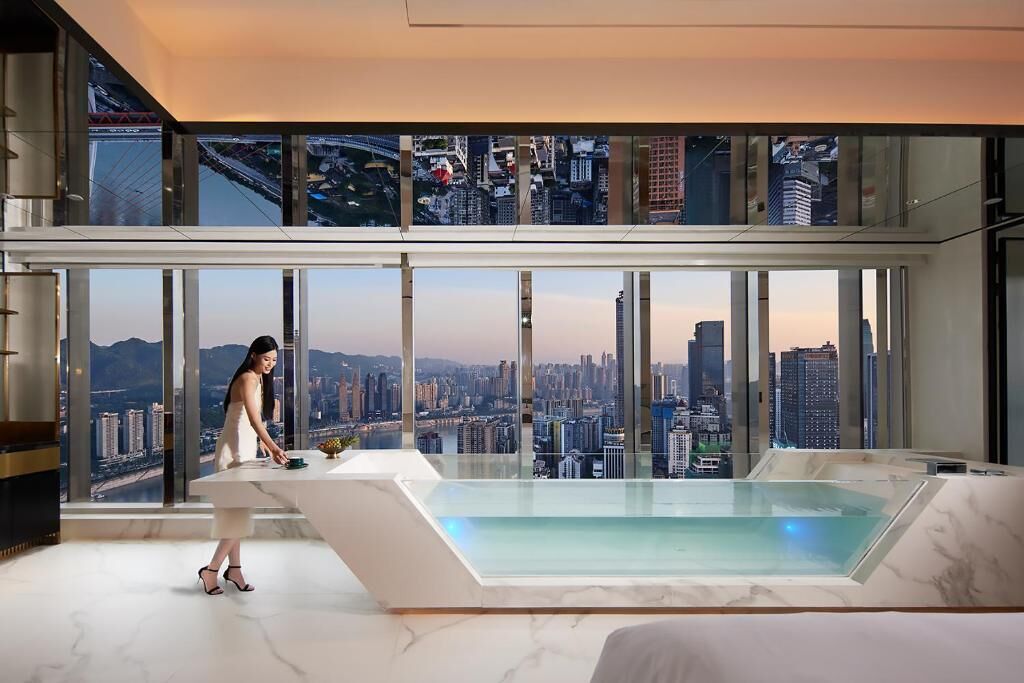 A modern hotel room with a large window offering a city view, a marble-topped desk, and a glass-enclosed hot tub.