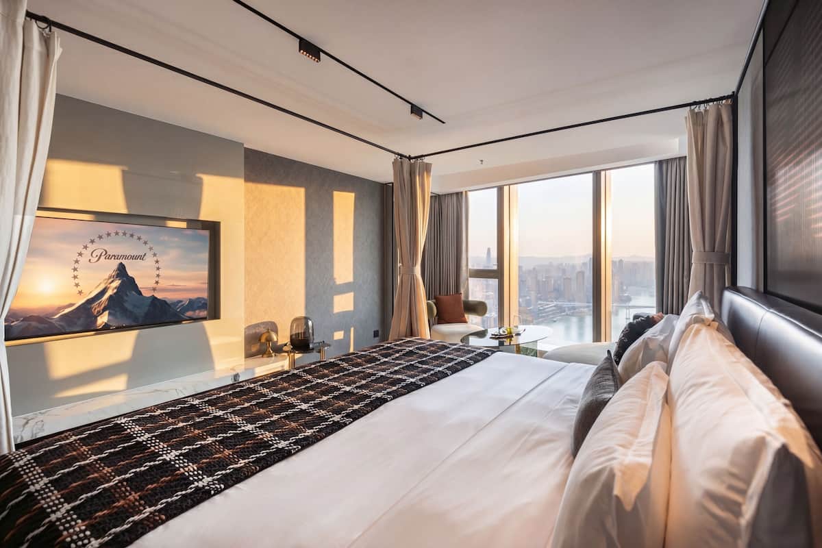 Ein Hotelzimmer mit einem großen Bett, Blick auf die Stadt, einem gerahmten Bild an der Wand und einem kleinen Tisch mit einer Lampe.