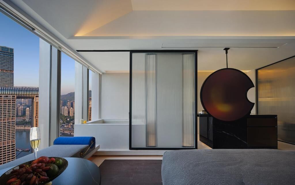 A modern hotel room with a large window offering a city view, a sliding glass door, and a minimalist design.