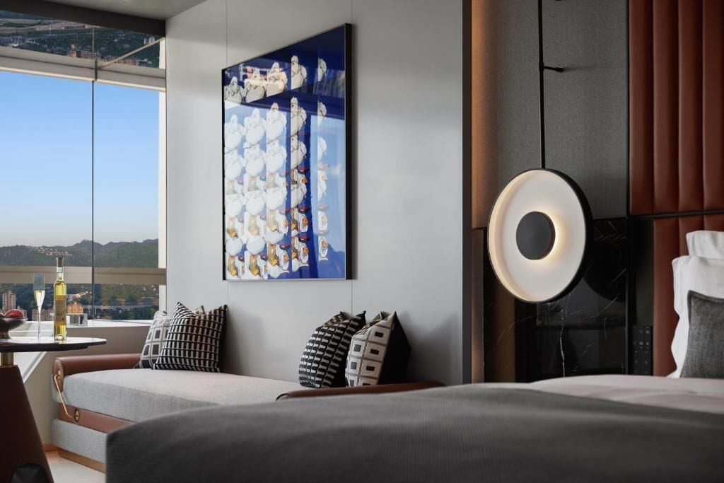 A modern hotel room with a large window, a painting on the wall, a round table with a bottle, and a bed with pillows.