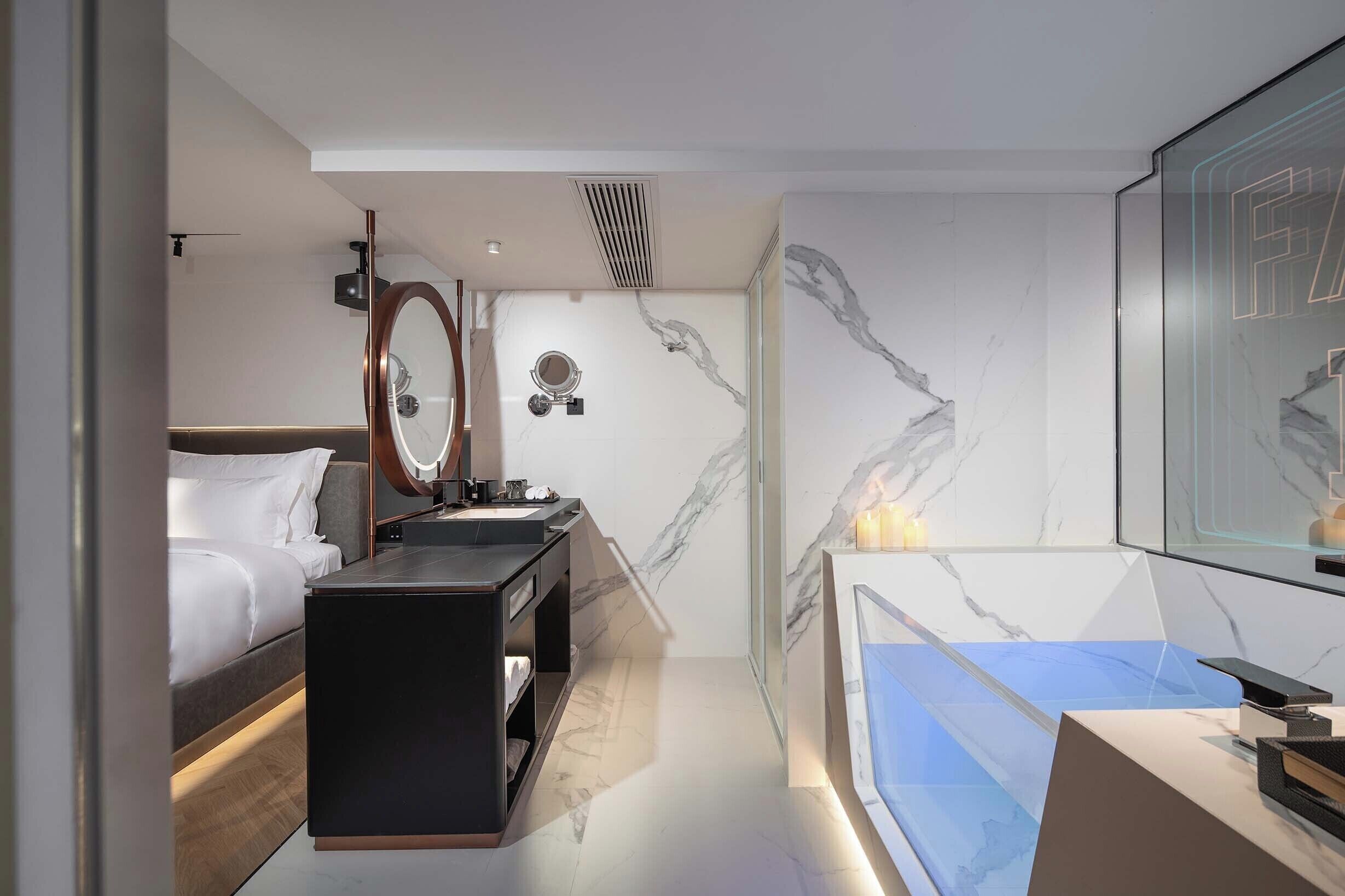 A modern hotel room with a bed, a vanity with a mirror, a bathtub, and marble walls.