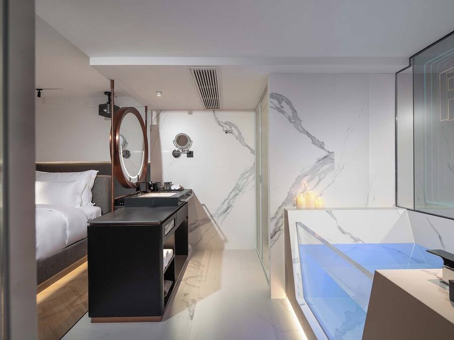A modern hotel room with a bed, a vanity with a mirror, a bathtub, and marble walls.