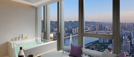 A modern hotel room with a large window offering a city view, a small sitting area with a magazine, and a bottle of wine.
